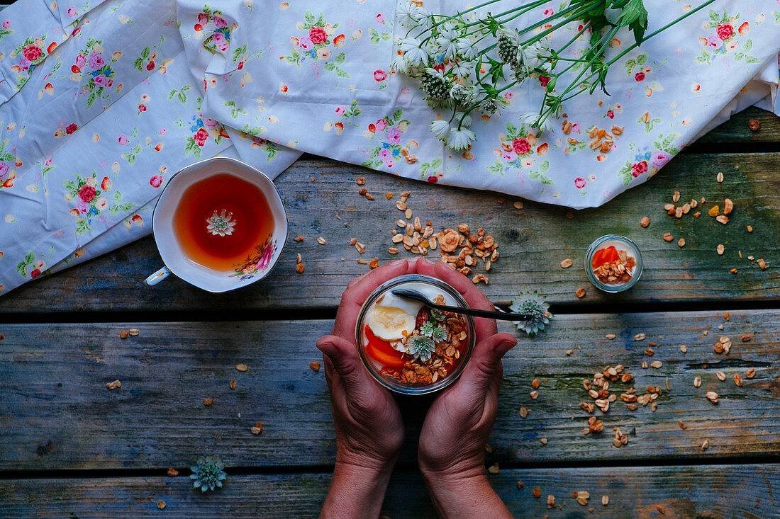 A jar with yoghurt, granola and fresh fruit with two hands on a wooden table with a cup of tea and flowers