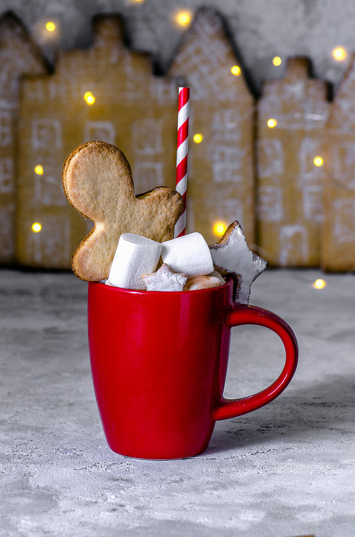 Red cup with cocoa, marshmelow, gingerbread man, star and a straw in a strip on the background of a gingerbread town. Christmas style
