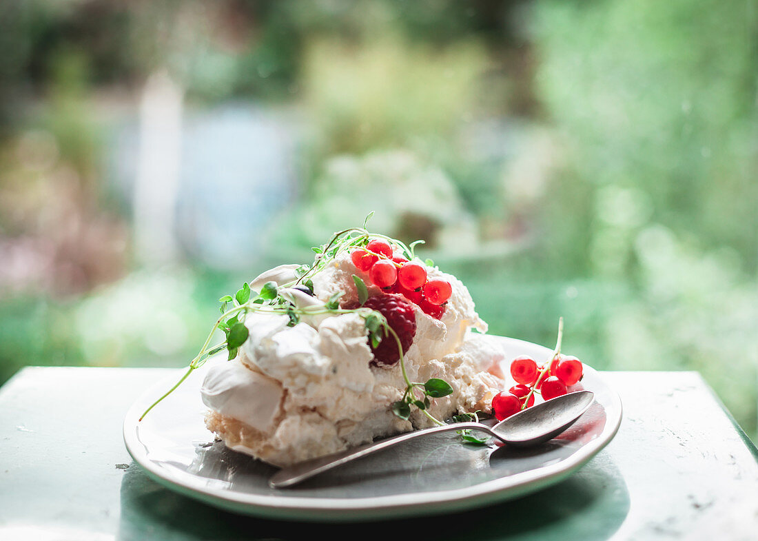 A slice of Pavlova topped with redcurrants and raspberries, decorated with thyme