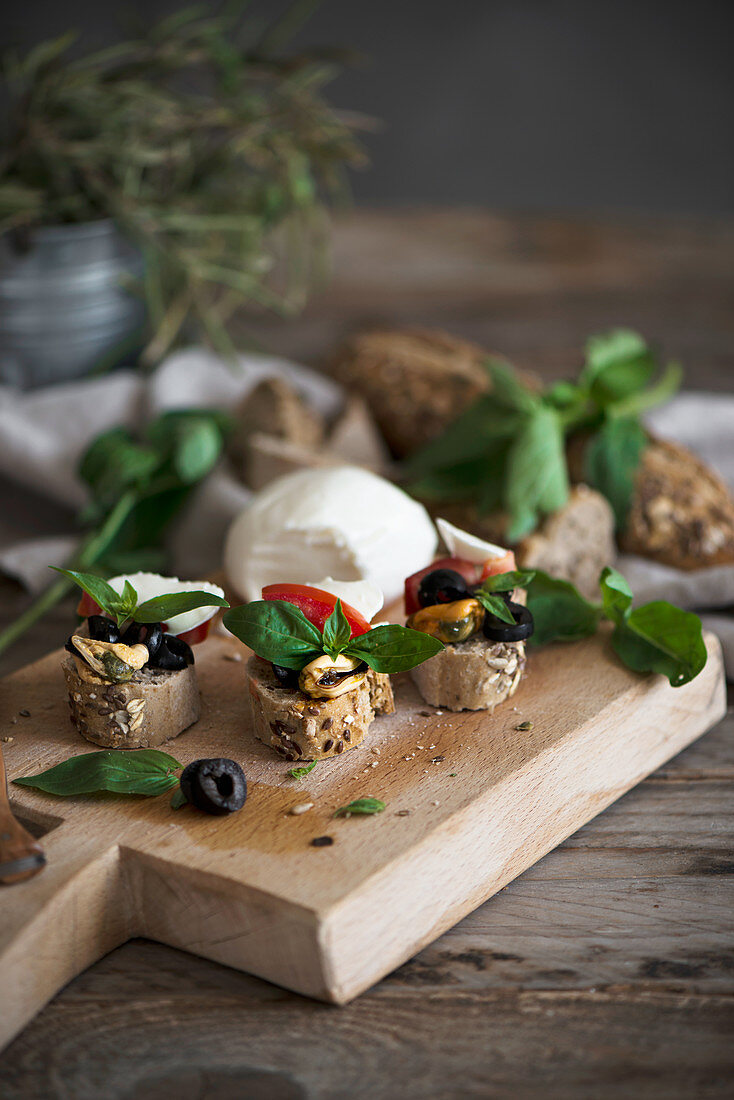 Crostini with mozzarella, olives, mussels, tomatoes, and basil leaves on a wooden board