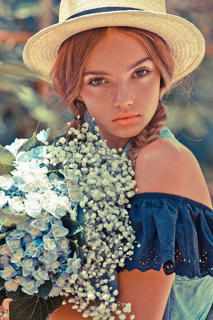 A young woman wearing a straw hat and a blue off-the-shoulder blouse with a bunch of flowers