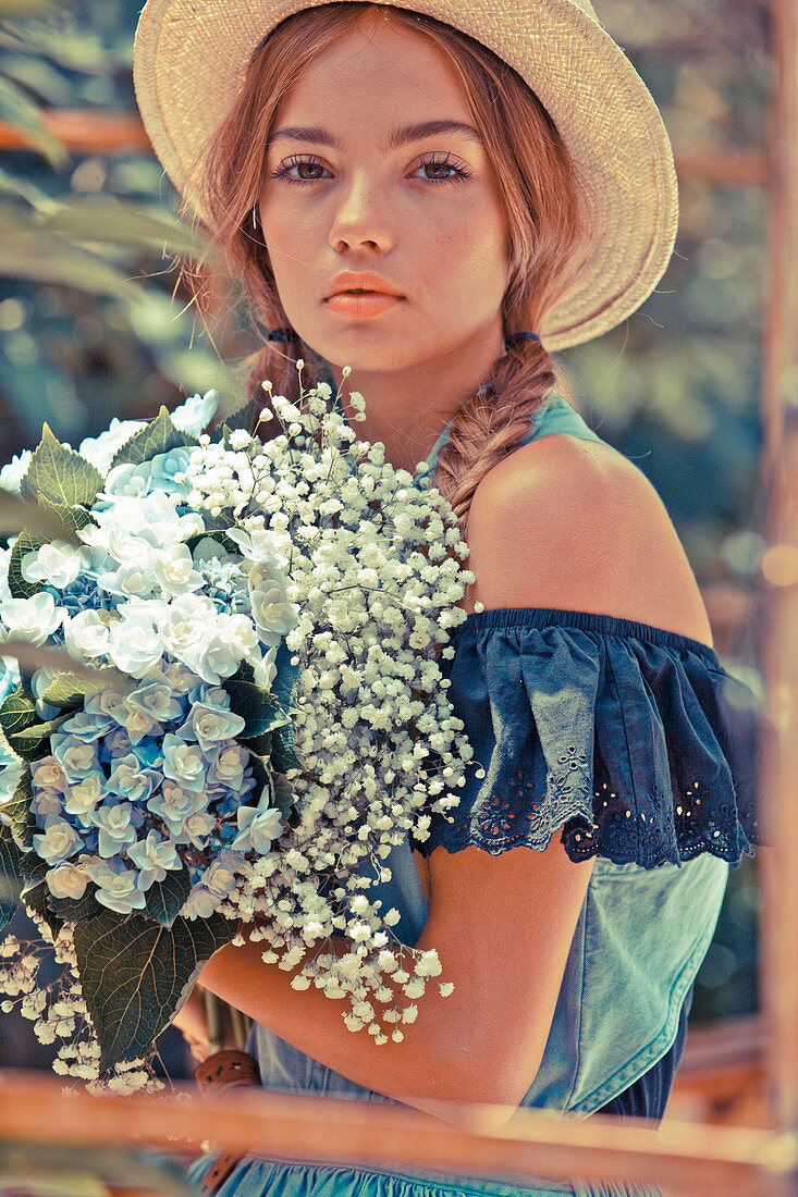 A young woman wearing a straw hat and a blue off-the-shoulder blouse with a bunch of flowers