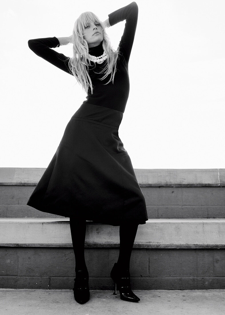 A blonde woman wearing a black roll neck jumper, a black skirt and tights (black-and-white shot)