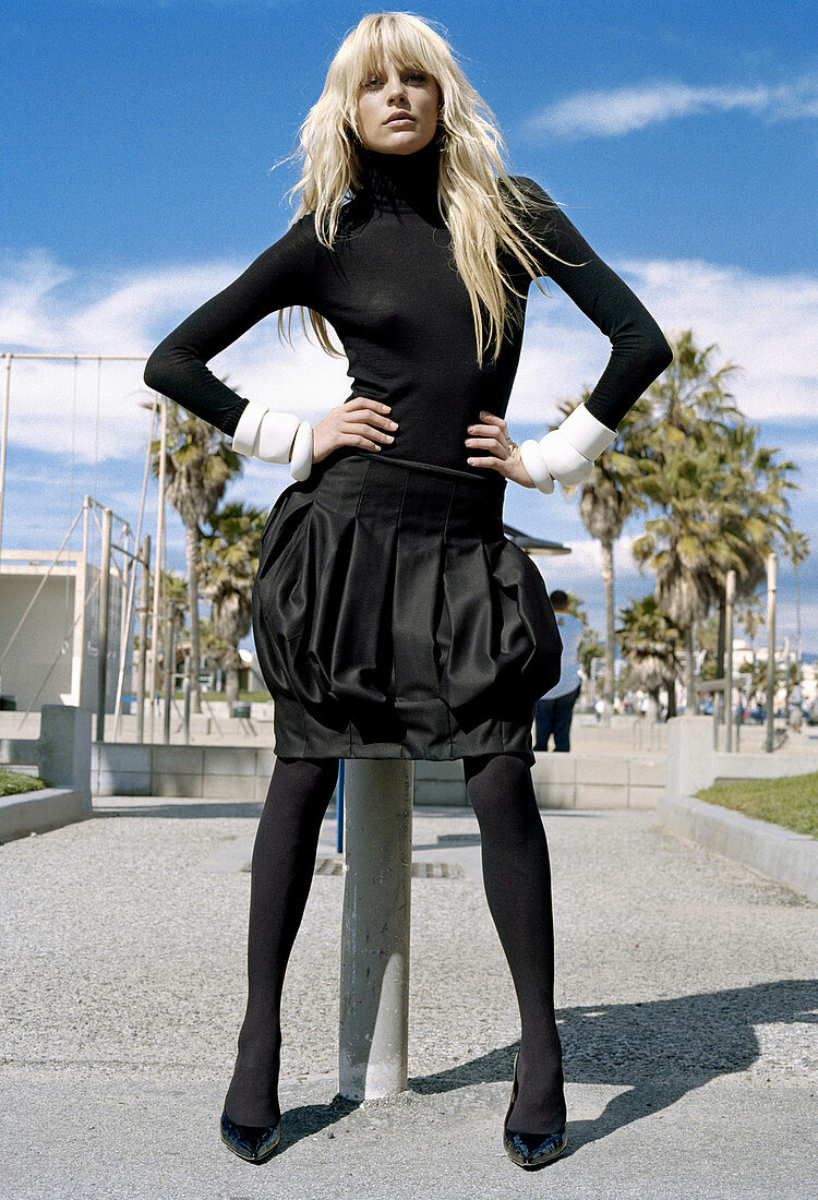 A blonde woman wearing a black turtle neck jumper, a black skirt and tights