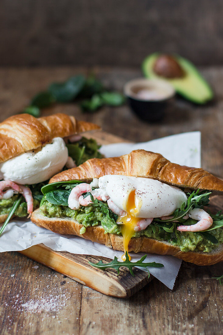 Croissant with avocado puree, shrimps, poached egg, rocket and spinach, salt in a bowl, avocado, spinach leaves