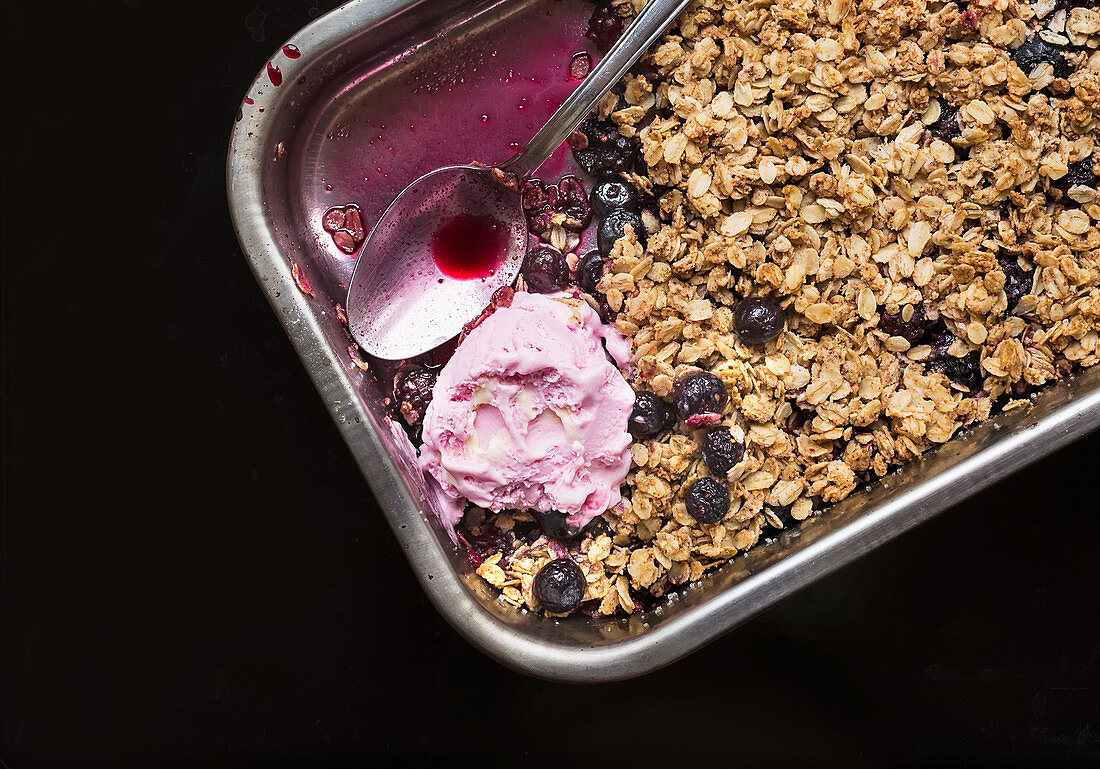 Oat granola with fresh berries and strawberry ice-cream in a silver baking dish over a black background