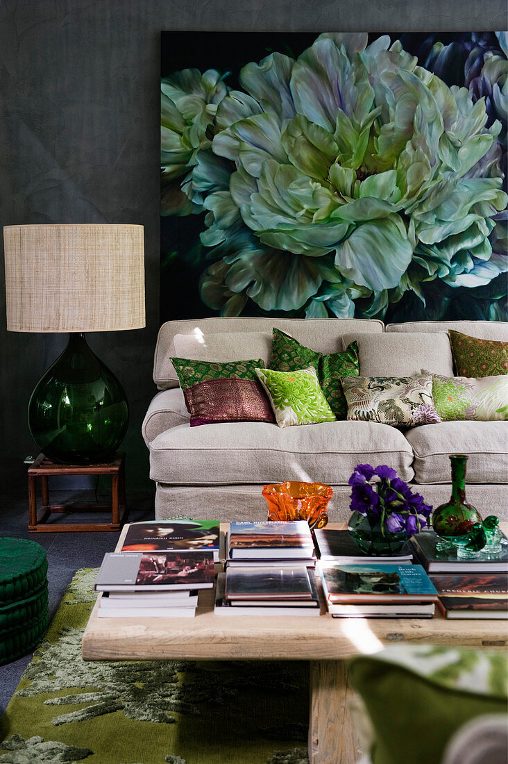 View of books on wooden table and sofa with scatter cushions below large painting on wall