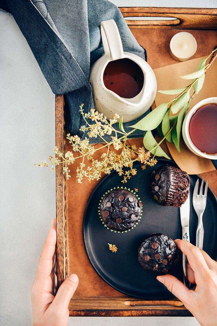 Person holding a wooden tray with healthy chocolate zucchini muffins on a black plate sand tea