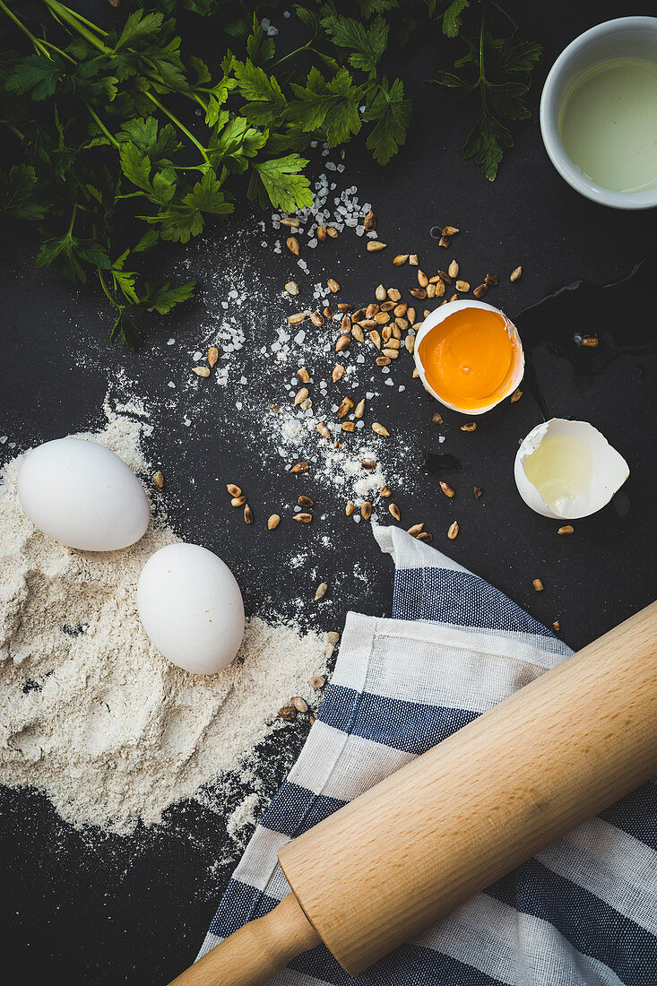 An arrangement of ingredients and utensils: eggs, flour, herbs, pine nuts and a rolling pin