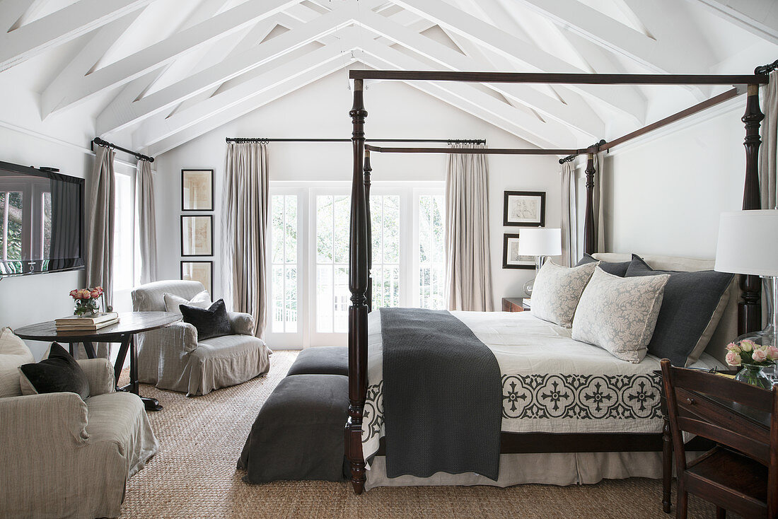 Four-poster bed, loose-covered armchairs and round table in country-house bedroom with exposed wooden roof structure
