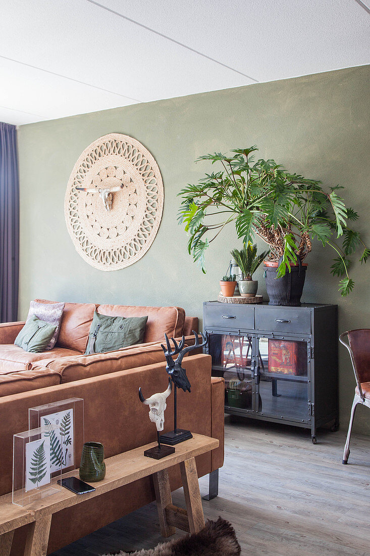 Ledersofas, schwarzes Schränkchen mit Grünpflanzen und Wandteppich mit Tierschädel im Wohnzimmer mit grüner Wand