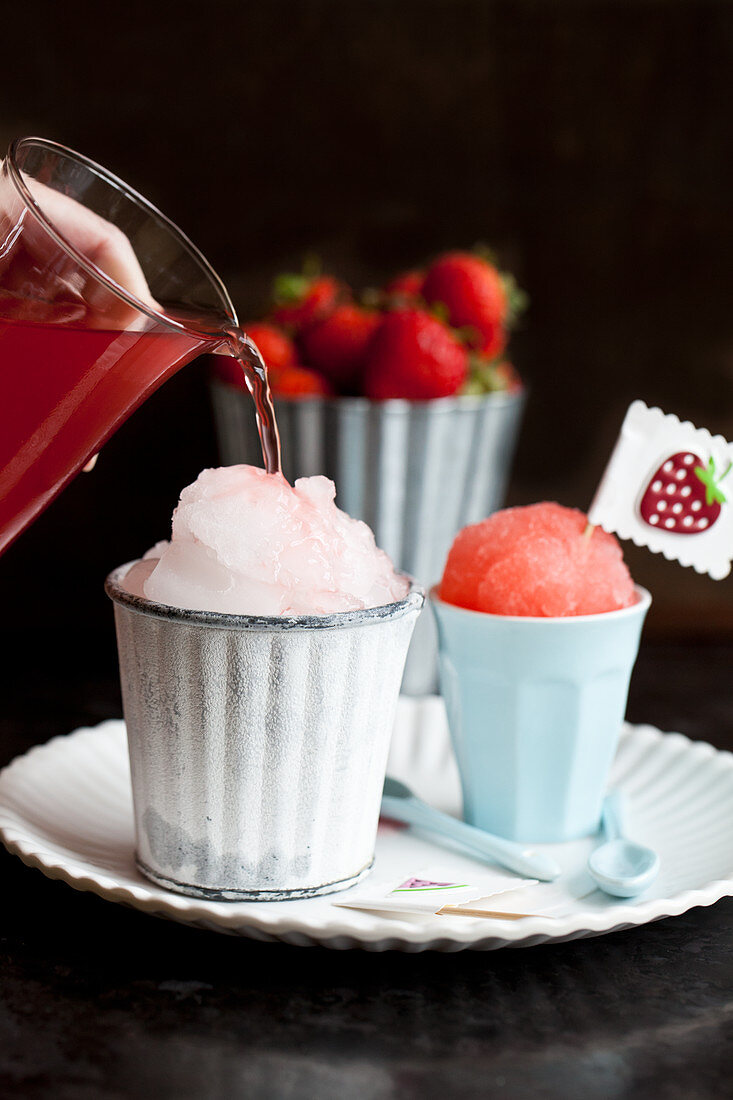 Homemade snocones in tiny cups with strawberry syrup being poured onto crushed ice, strawberries and fruit stickers on flags