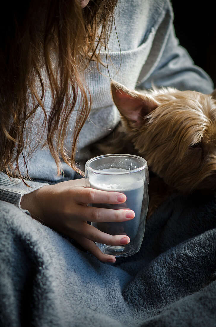 Frau sitzt entspannt mit Hund und Aktivkohle-Latte in Decke gehüllt