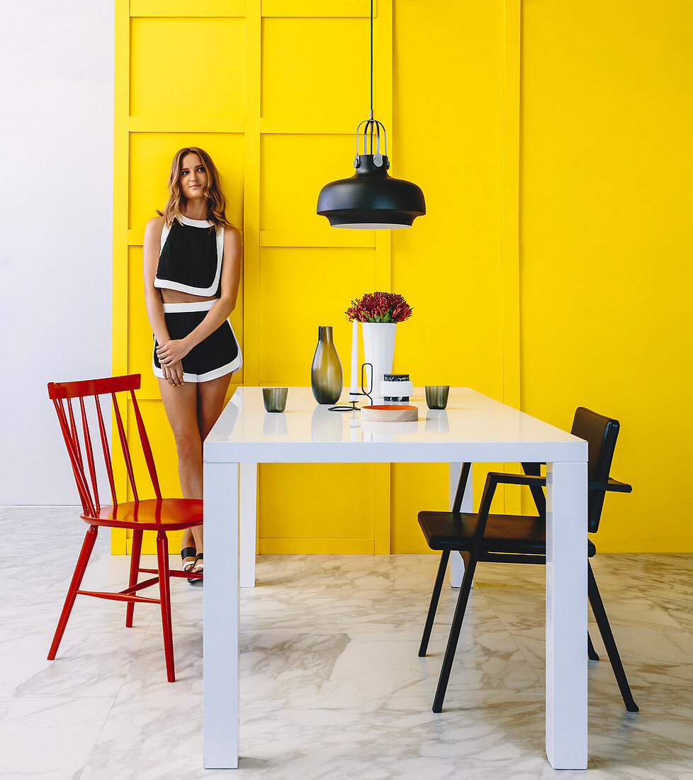 Weißer Tisch mit Stühlen im Essbereich, junge Frau vor gelber Kassettenwand