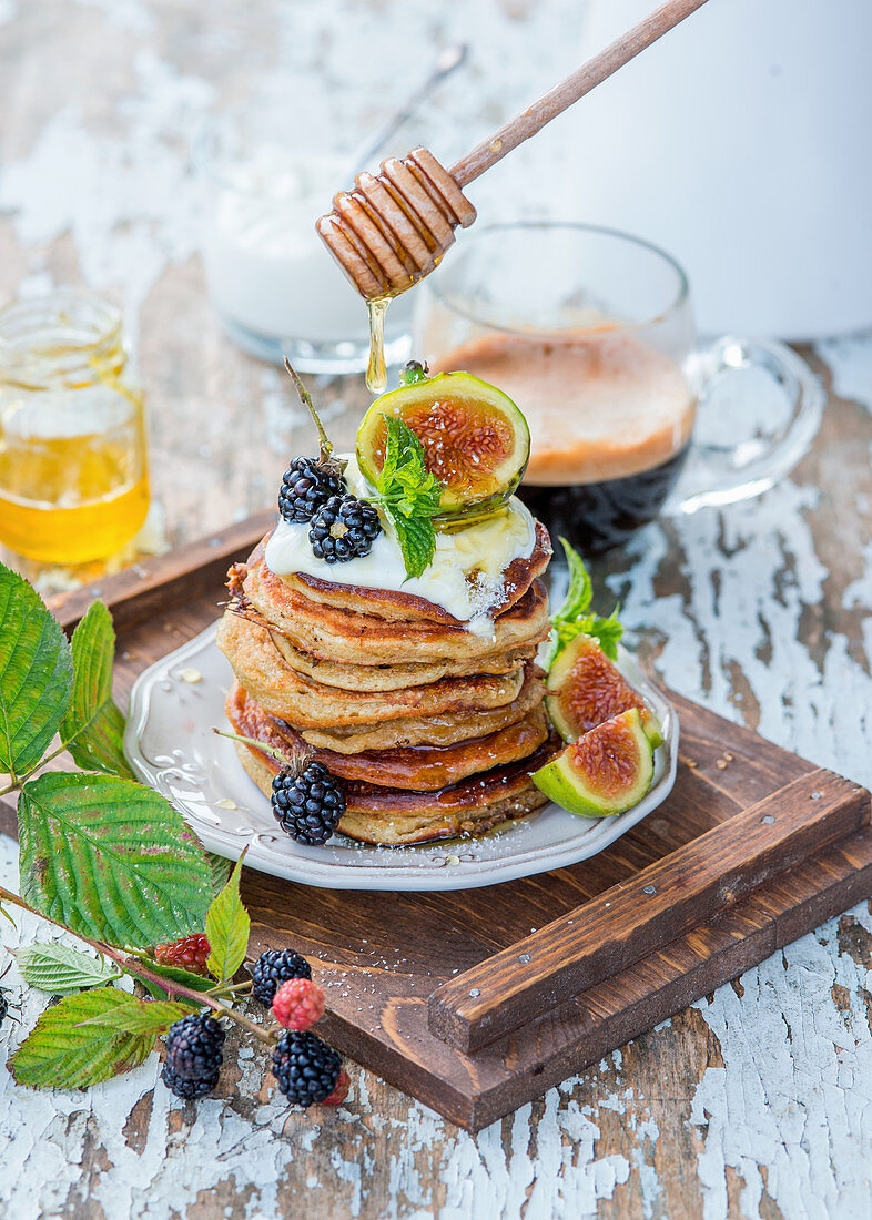 Honey being drizzled over a stack of pancakes with yoghurt, figs and blackberries