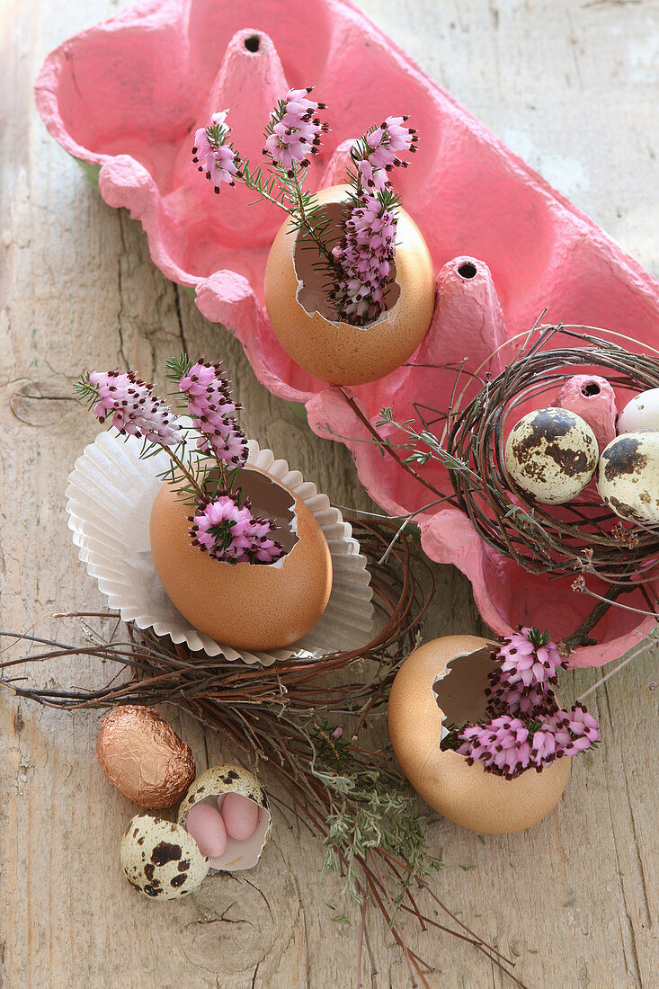 Ausgepustete Eier als kleine Blumenvasen im rosa Eierkarton mit lila Heidekrautblüten und Zuckereiern