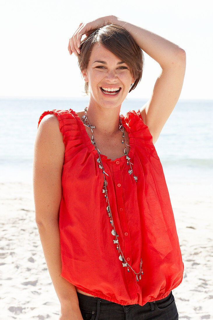 Reife brünette Frau mit rotem Shirt, Halskette und schwarzer Shorts am Strand