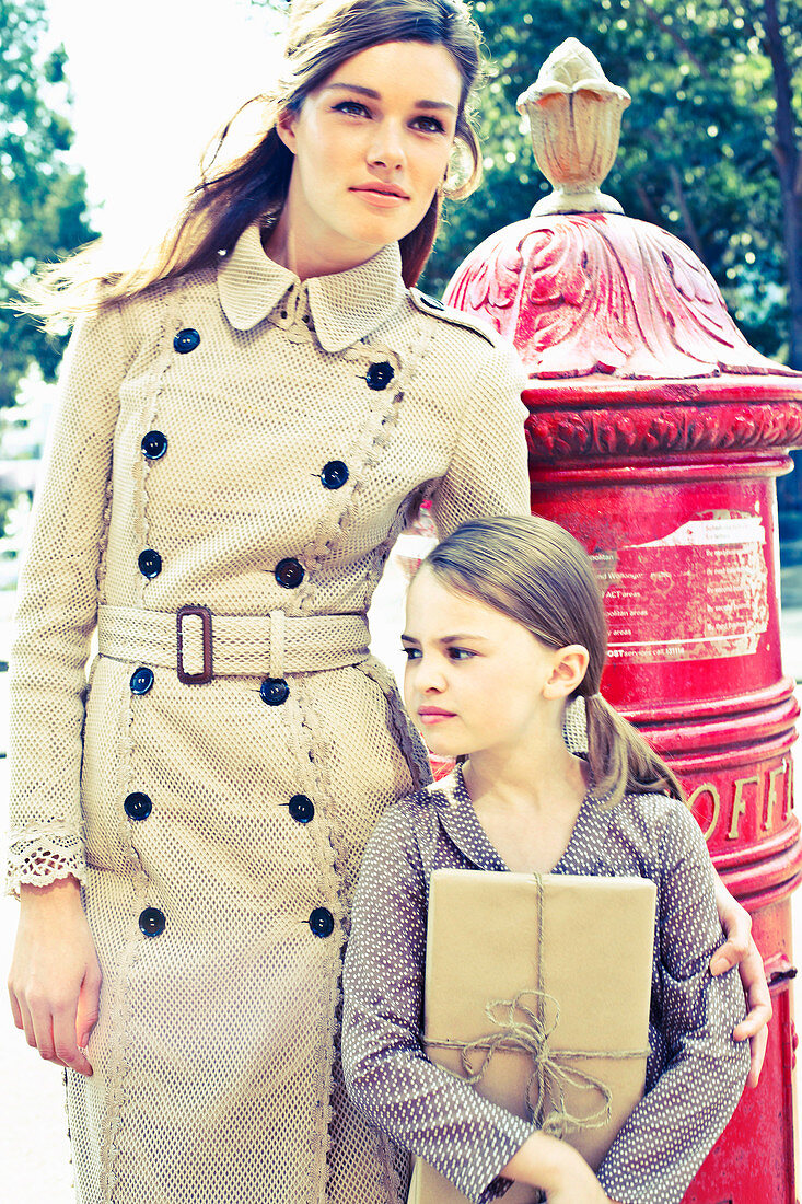 Junge Fau im beigen Mantel und Mädchen mit Packet stehen neben einem englischen Briefkasten
