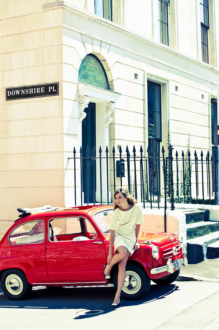 A young brunette woman wearing a summer dress leaning against a car