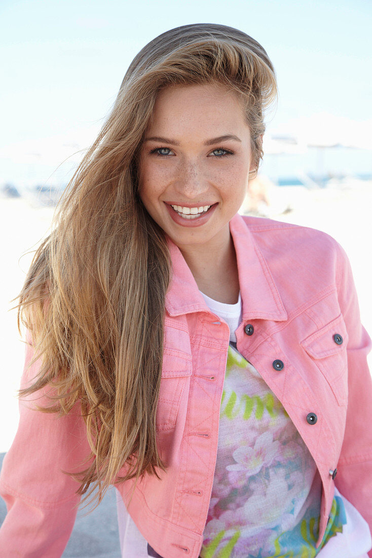 A young blonde woman on a beach wearing a printed t-shirt and a pink denim jacket