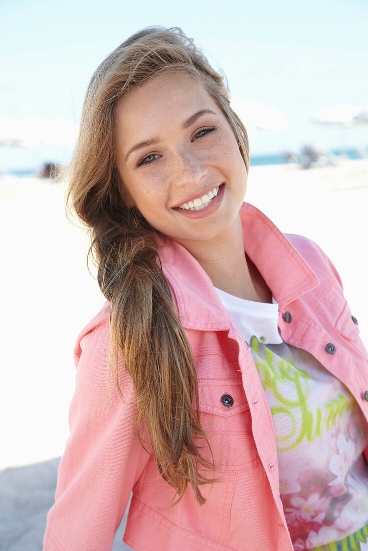 A young blonde woman on a beach wearing a printed t-shirt and a pink denim jacket