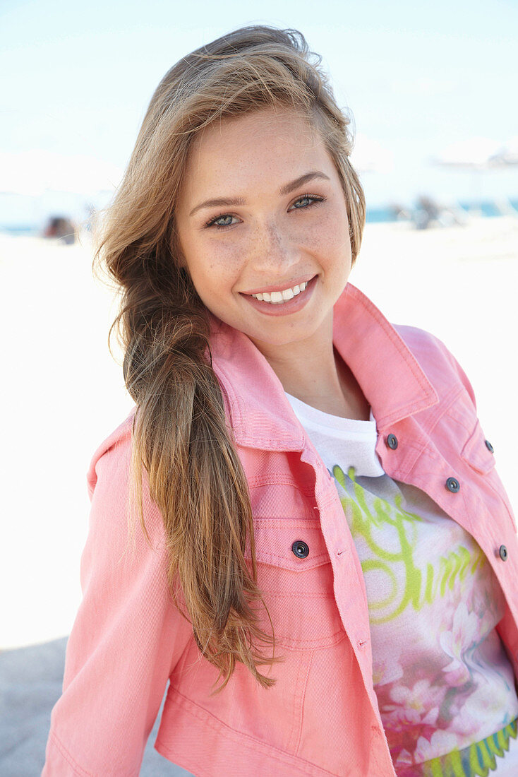 A young blonde woman on a beach wearing a printed t-shirt and a pink denim jacket