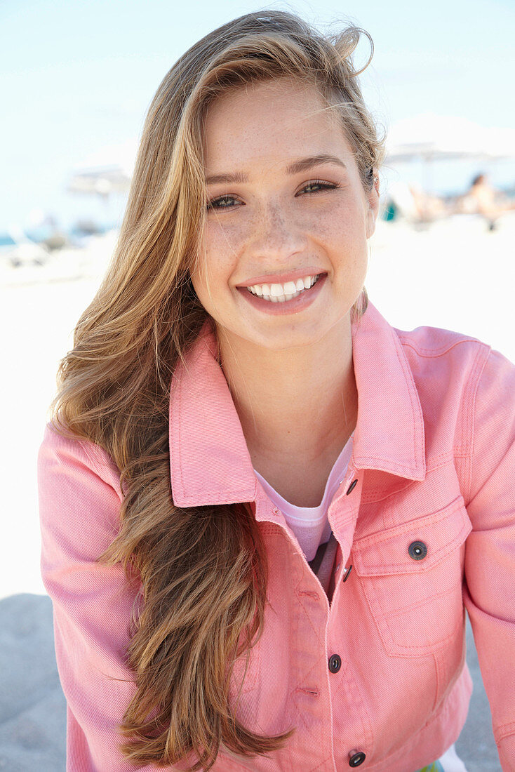 A young blonde woman on a beach wearing a t-shirt and a pink denim jacket