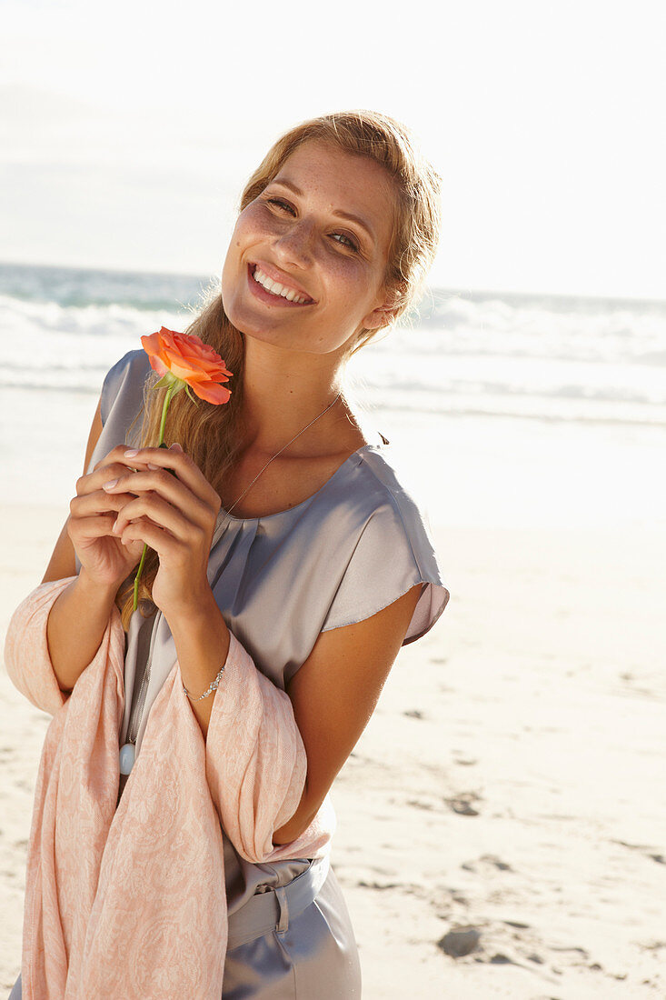 A mature blonde woman on a beach wearing a silver summer dress and holding an orange flower