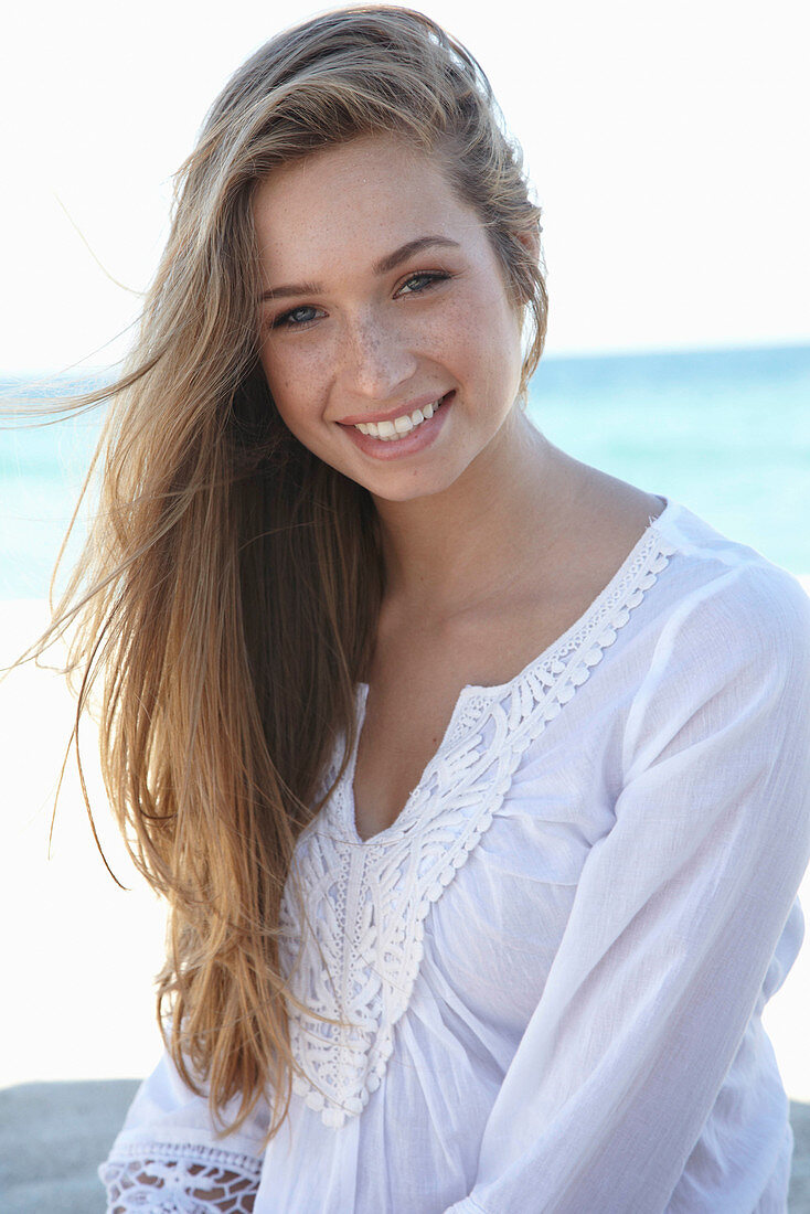 A young blonde woman on a beach wearing a white summer dress