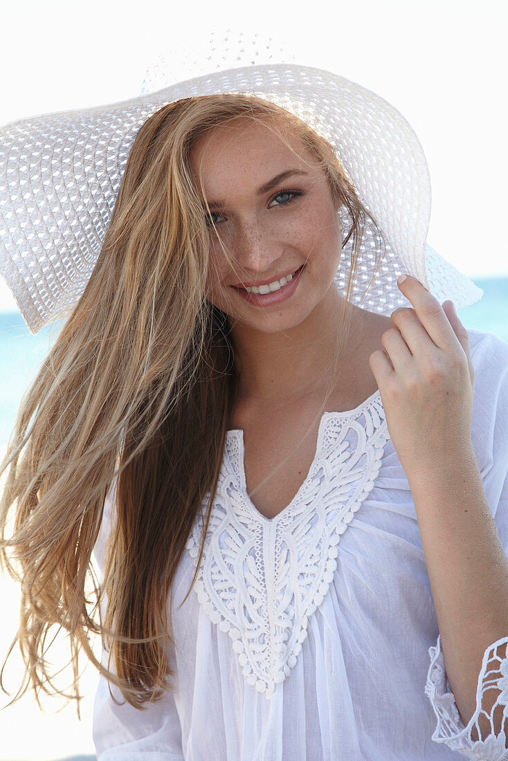 Junge blonde Frau mit weißem Sommerhut in weißem Sommerkleid am Strand