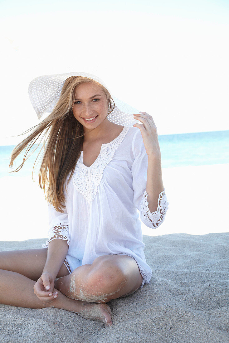 A young blonde woman on a beach wearing a white summer dress and a white summer hat