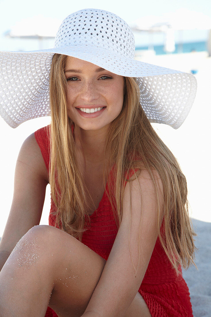 A young blonde woman on a beach wearing a red summer dress and a white hat