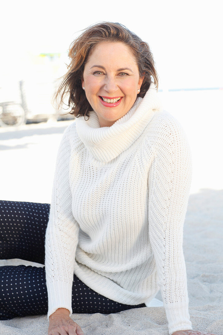 Brünette Frau in weißem Strickpullover und dunkler Hose am Strand