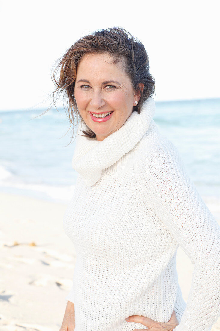 A brunette woman on a beach wearing a white knitted jumper