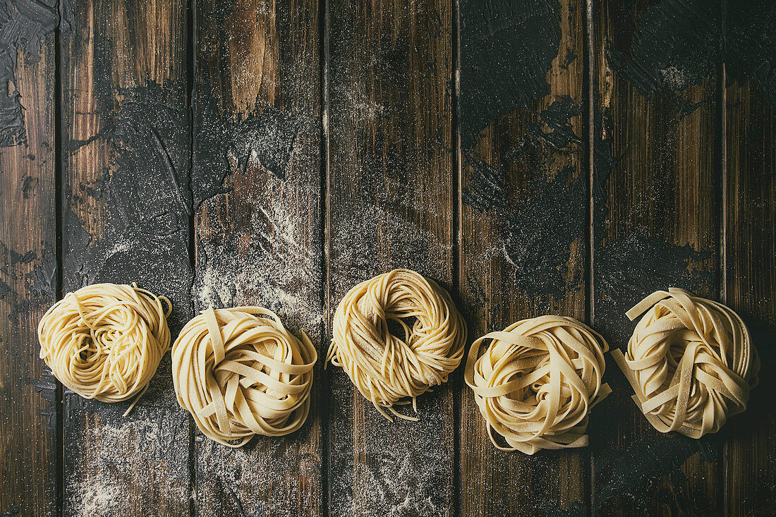 Variety of italian homemade raw uncooked pasta spaghetti and tagliatelle in row with semolina flour on wooden table