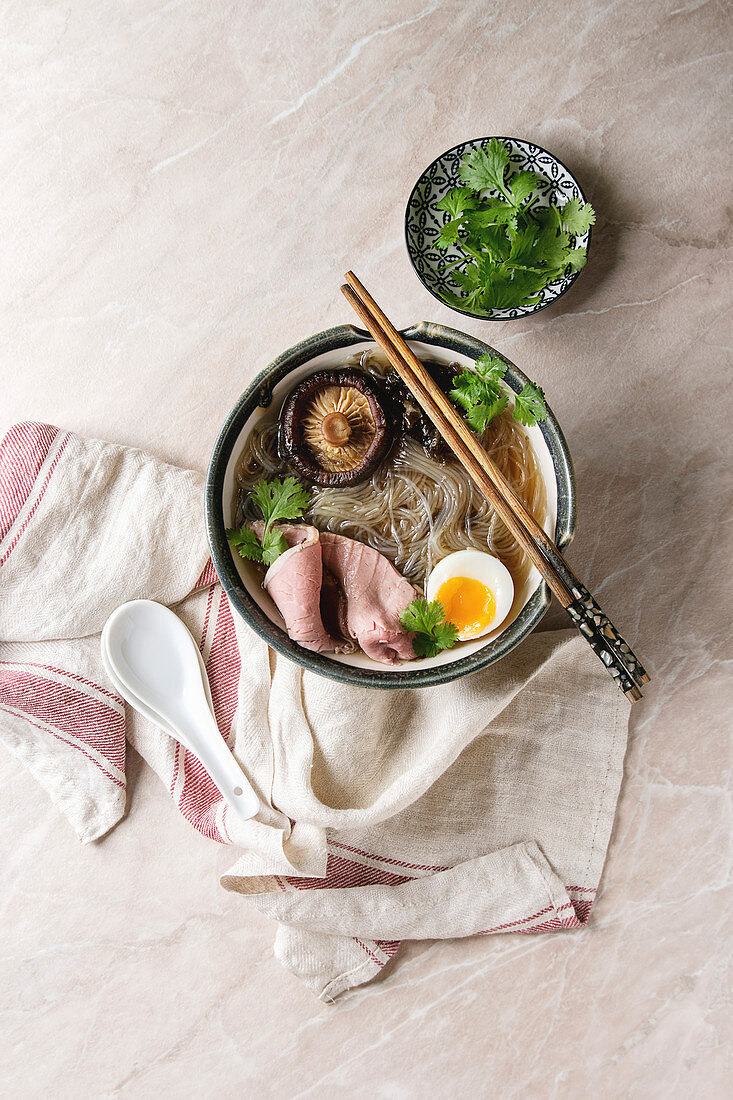 Traditional Japanese Noodle Soup with shiitake mushroom, egg, sliced beef and greens in ceramic bowl with wooden chopsticks
