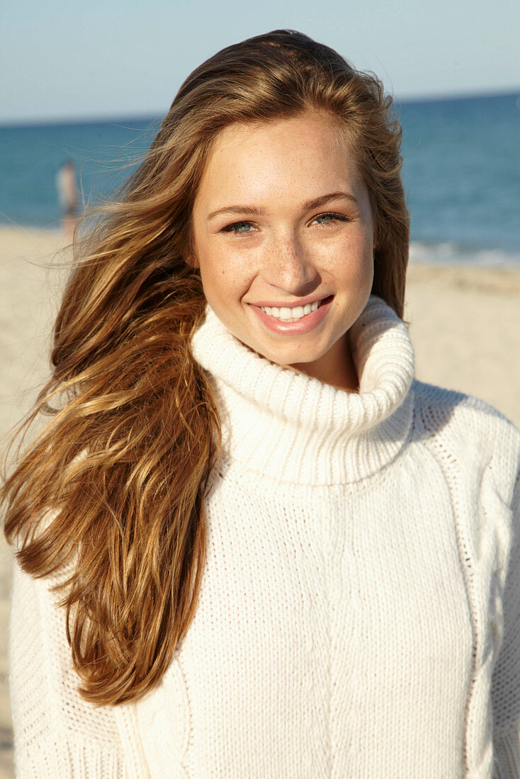 A young blonde woman on a beach wearing a white turtle neck jumper