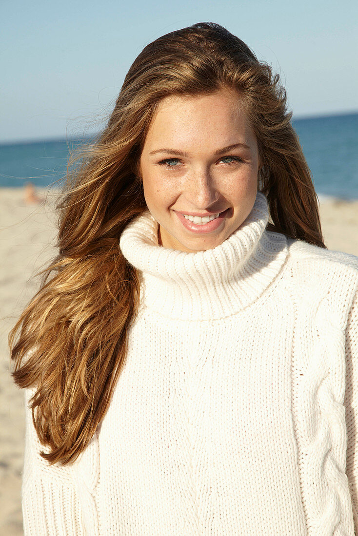 A young blonde woman on a beach wearing a white turtle neck jumper