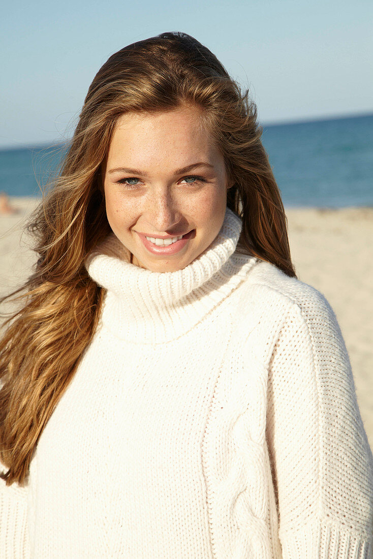 A young blonde woman on a beach wearing a white turtle neck jumper