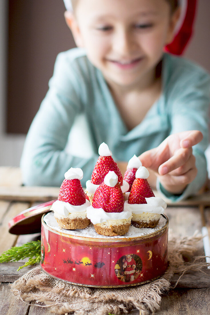Junge sitzt vor Mini-Käsekuchen in Keksdose dekoriert mit Weihnachtsmützen aus Erdbeeren