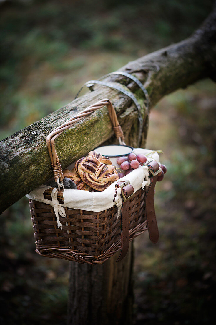 Picknickkörbchen, gefüllt mit veganem Schokoladenbrötchen, Obst und Kaffee, hängt an Holzgeländer