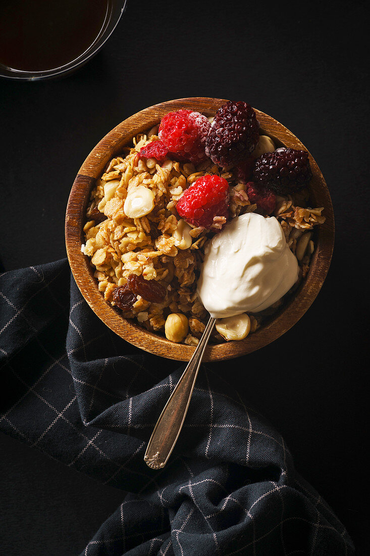Granola with raspberries and blackberries and a spoon of cream in wooden bowl with a cup of coffee on black table