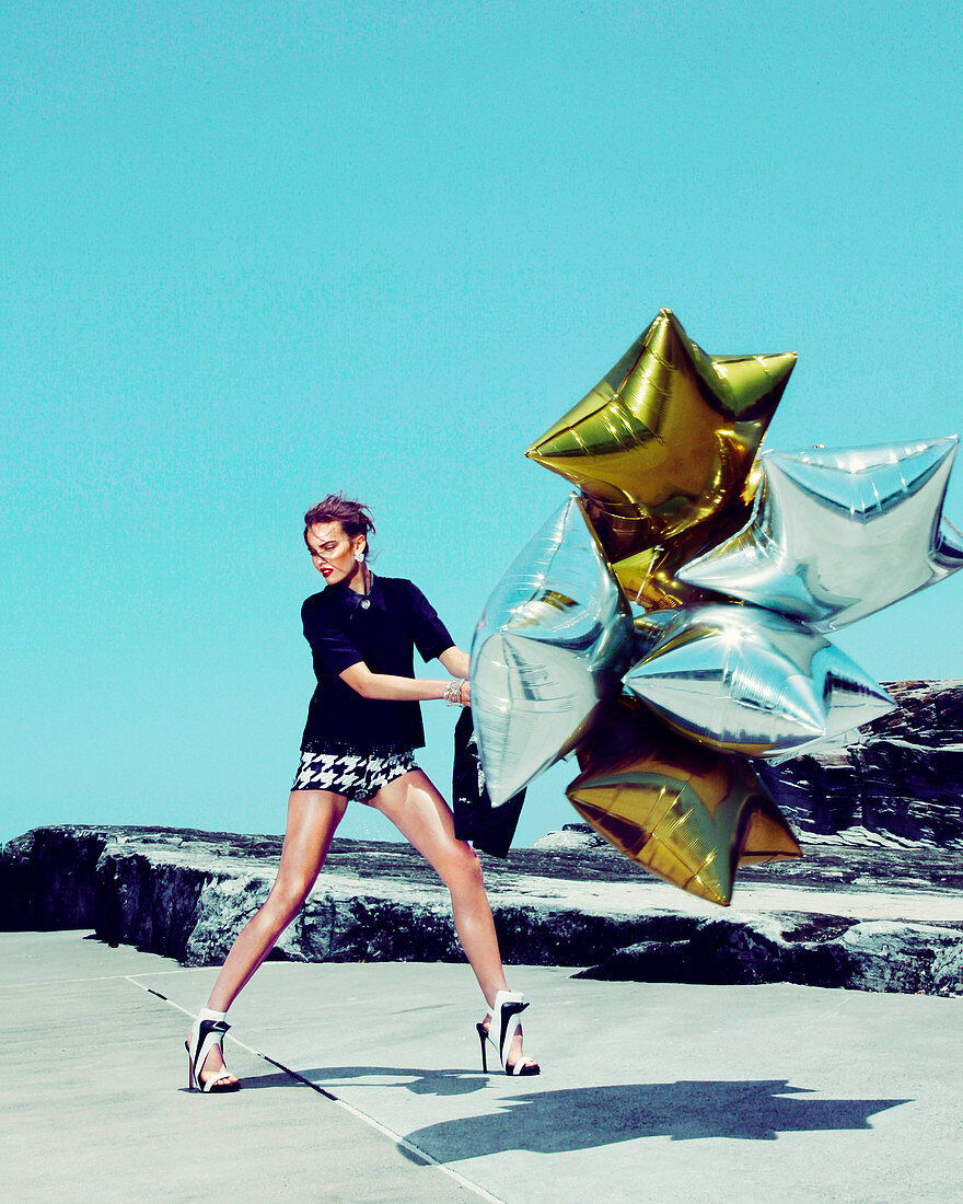 A young woman wearing a black shirt and patterned hot pants holding gold and silver balloons