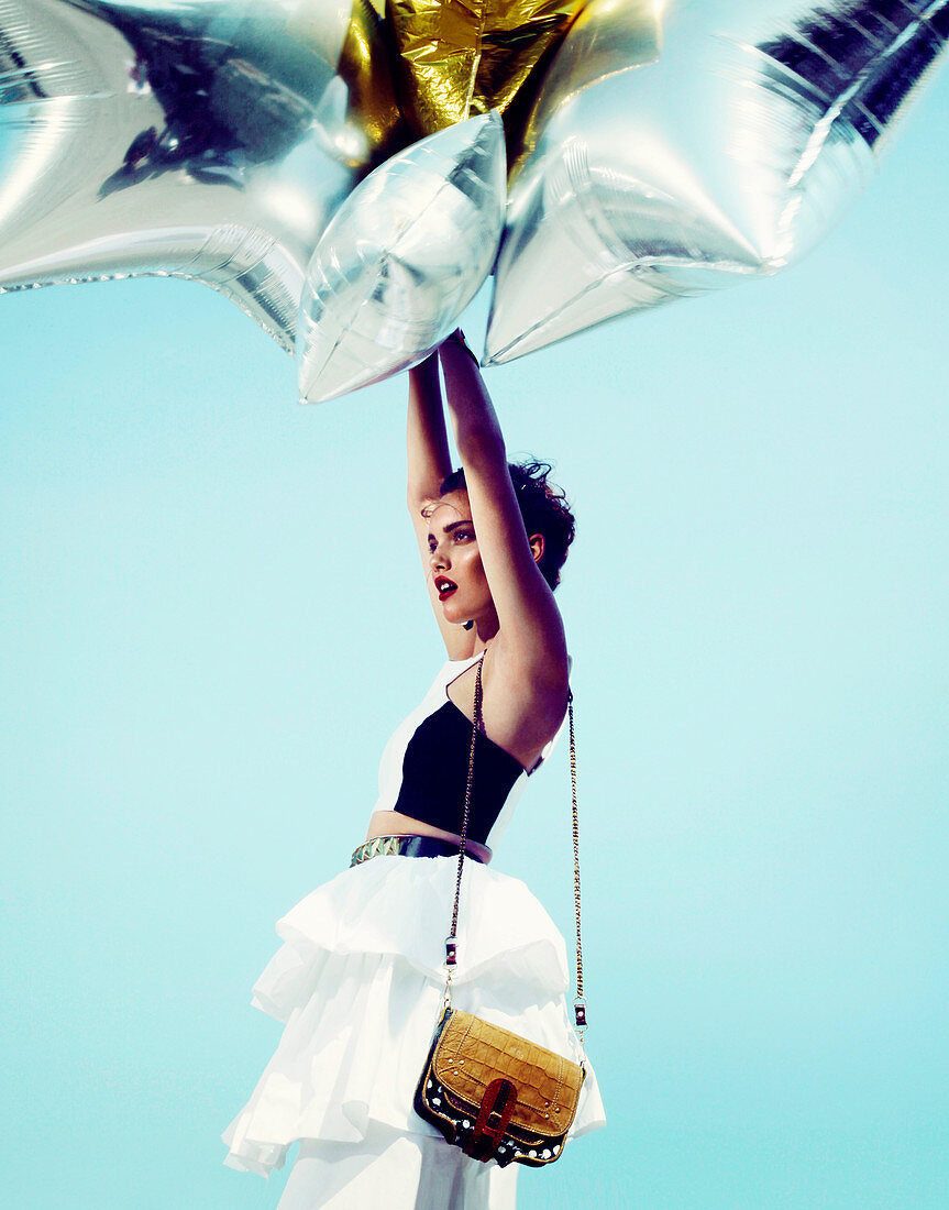 A young woman wearing a white skirt and a black-and-white top holding gold and silver balloons