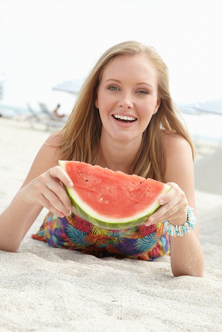 A young blonde woman on a beach wearing a colourful summer dress holding a wedge of melon