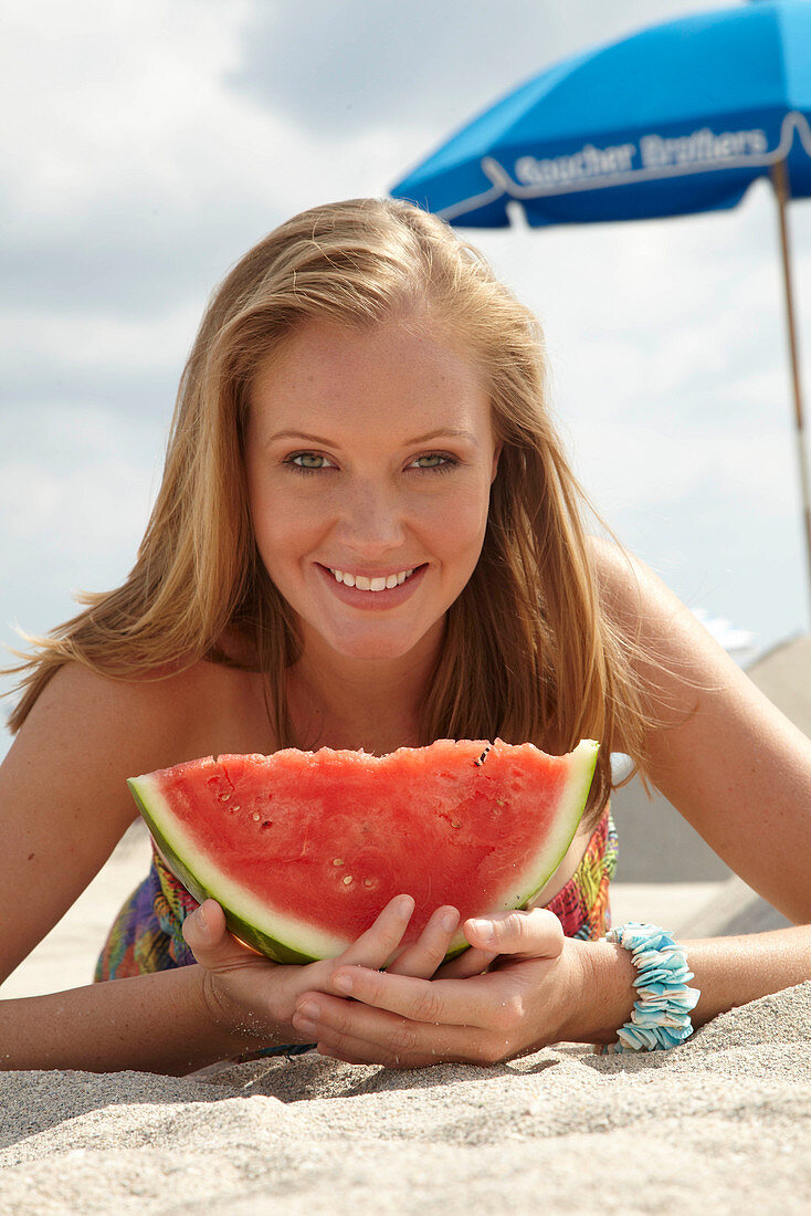 Junge blonde Frau im bunten Sommerkleid hält Melone am Strand