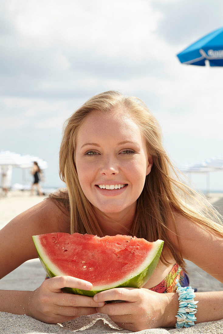 A young blonde woman on a beach wearing a colourful summer dress holding a wedge of melon