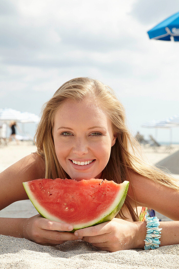 A young blonde woman on a beach wearing a colourful summer dress holding a wedge of melon