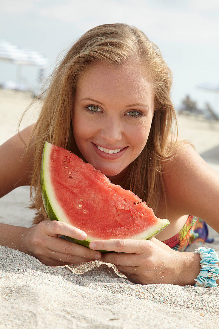 Junge blonde Frau im bunten Sommerkleid hält Melone am Strand