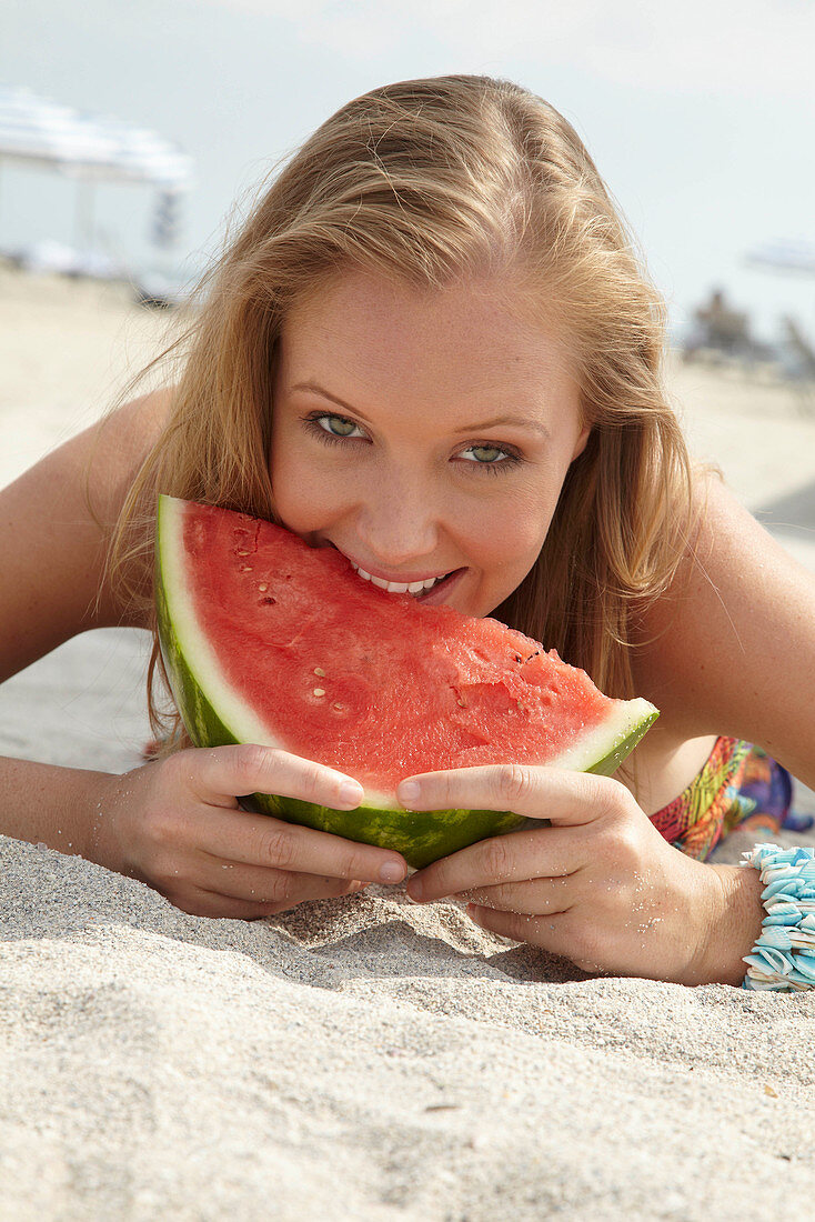 Junge blonde Frau im bunten Sommerkleid hält Melone am Strand